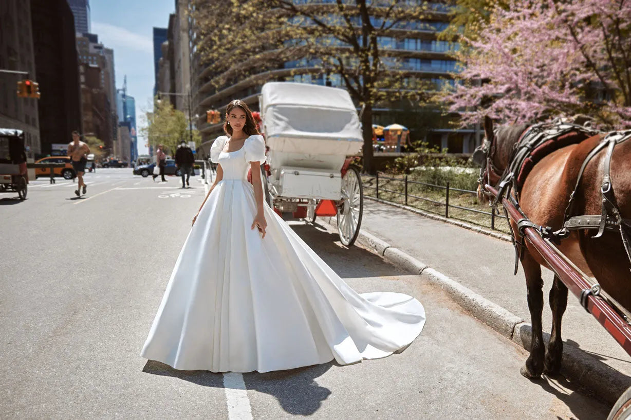 Gorgeous dress for a princess wedding look. The lush gown is made of glossy fabric and has no additional decorations. The corset is embellished with delicate Boat neckline and elbow-length loose sleeves with take-ups in the shoulders zone.